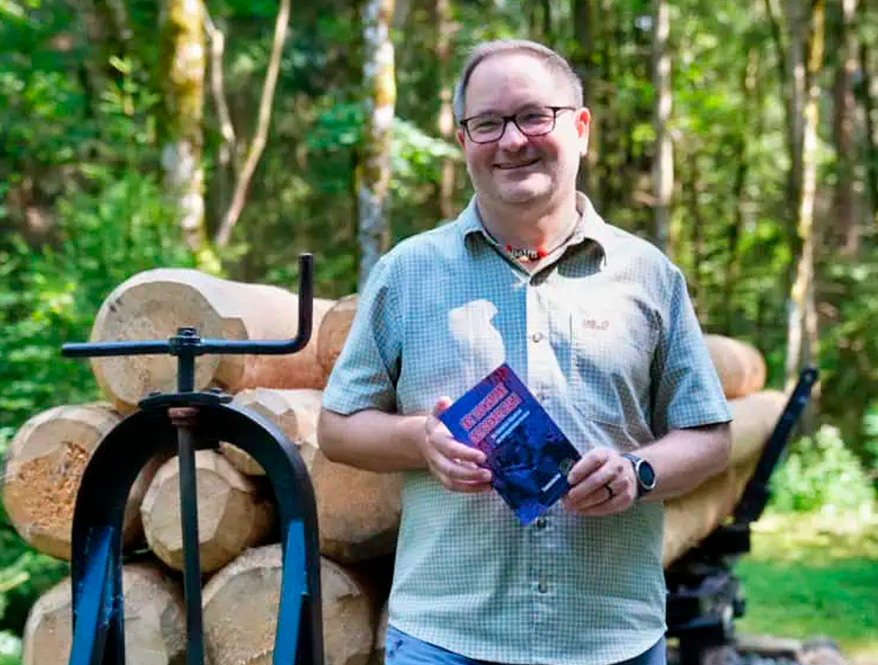 Auf dem Bild ist Thomas Kockler, der Autor des Buches "Der Tod kam mit der Bockerlbahn" zu sehen. Auf dem Bild trägt er eine Brille und ein kariertes beiges Hemd. Er hält sein Buch in den Händen. Hinter ihm liegen mehrere Holzstämme auf einem Wagen, wie er vor ca. 100 Jahren für den Holztransport genutzt wurde. Auf dem Foto steht er in einer waldreichen Umgebung, die in Verbindung mit der historischen Bockerlbahn steht – der alten Trasse der Bockerlbahn, die heute als Wanderweg zu begehen ist.